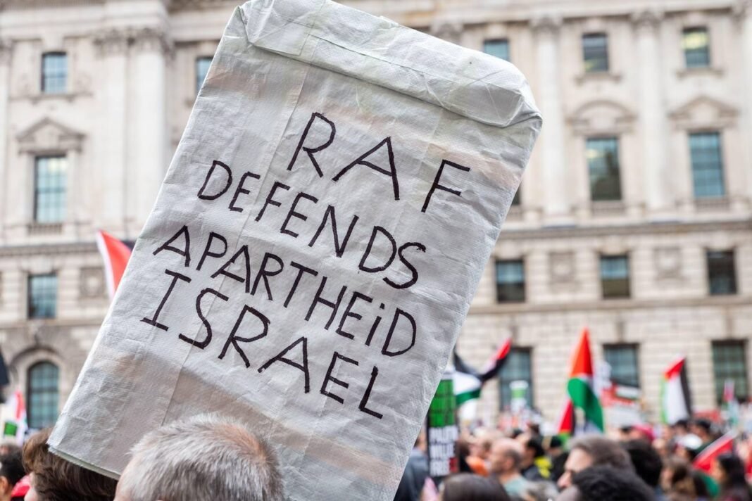 Pro Palestine protesters protesting in London, UK, with a placard referencing military support for Israel.