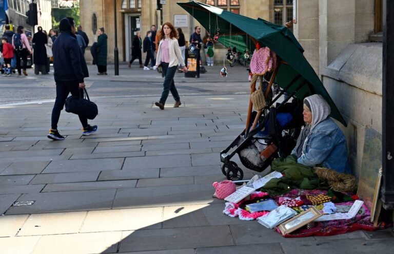 Older woman bundled up selling items while sitting on pavement in Oxford, UK